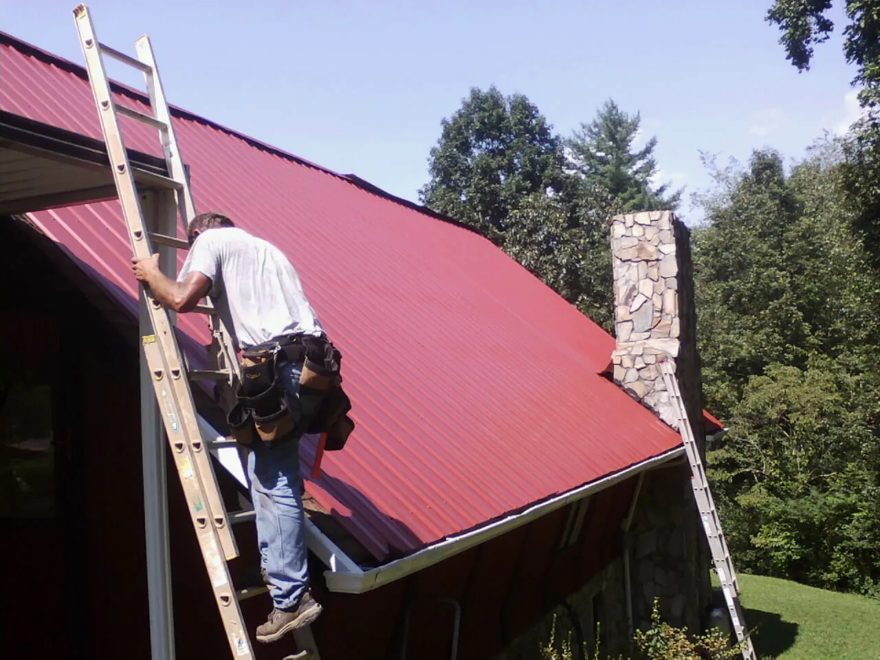 Roofer on a ladder beside a red metal roof and stone chimney — Soffit & Fascia Repair by Roofing Brothers in Cooke City